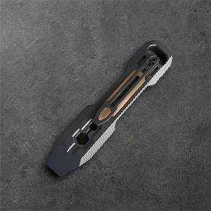 May include: A multi-functional metal pry bar tool featuring a matte black finish with silver-toned textured side grips and a central bronze-colored inlay. The tool includes a hexagonal cutout and a black pocket clip secured by two small screws.