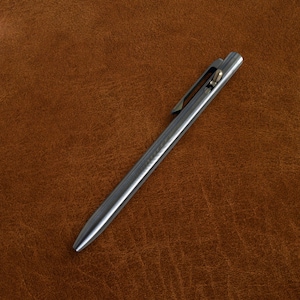 May include: A sleek, silver-colored pen with a clip and a pointed tip. The pen is lying on a brown leather surface. The pen's design is minimalist and modern, with a focus on functionality and style.