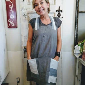 Könnte beinhalten: Eine Frau trägt eine graue Leinenschürze mit weißen und grauen Patchworktaschen. Die Schürze hat breite Träger und ein schlichtes Design.