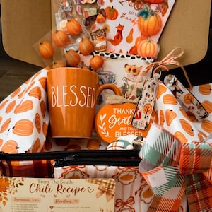 May include: A gift box containing an orange mug with the word BLESSED, a round sign reading THANKFUL AND TRULY BLESSED, a chili recipe card, pumpkin-themed wrapping paper, a plaid bow, and a sheet of fall-themed stickers featuring pumpkins and foxes.