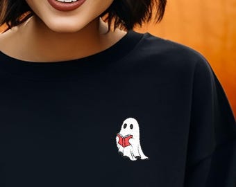 Puede incluir: Sudadera negra con un pequeño fantasma bordado leyendo un libro. El fantasma es blanco y el libro es rojo. La sudadera es de color negro liso. En el fondo hay varias calabazas naranjas grandes.