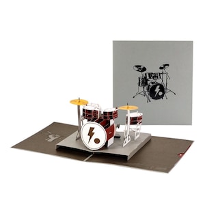 May include: A three-dimensional pop-up greeting card featuring a miniature drum kit in red, white, and black. The bass drum displays a white lightning bolt icon. A matching grey envelope is positioned behind the card, showing a black line drawing of a drum set.