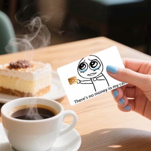 K&ouml;nnte beinhalten: Eine wei&szlig;e rechteckige Kreditkarte mit einer schwarz-wei&szlig;en Strichzeichnung einer weinenden Figur, die einen goldenen Chip h&auml;lt. Der Text "There's no money in my card" ist in Schwarz auf der Unterseite der Karte aufgedruckt.
