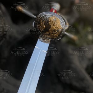 以下が含まれることがあります： 金色の柄と丸い装飾的なガードが付いた銀の剣。ガードには、馬に乗った騎士の浮き彫りの画像が描かれている。剣は暗い背景に飾られている。