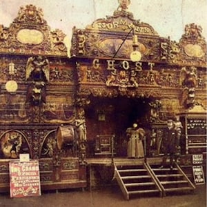 May include: Vintage photograph of a carnival attraction, "Norman's Ghost," with ornate wooden carvings and the word "GHOST" prominently displayed. A sign advertises "The Grand Spectacle." The image is sepia-toned.