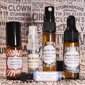 May include: Five small glass bottles with labels and black caps. The labels are colorful and feature circus-themed text and images. The bottles contain different scents, including Bourbon Spice Cake, The Bee's Knee, Queen's Pistachio Cream, She Crawled from the Swamp, and The Snow Queen's Snowflakes.