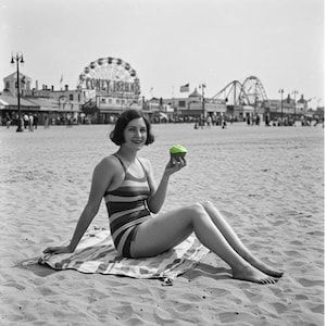 May include: A woman in a striped swimsuit sits on a beach towel, holding a green cupcake. She is smiling and looking at the camera. In the background, there is a Ferris wheel and other amusement park rides.