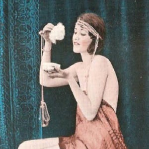 May include: A woman in a pink dress with a white headband is applying makeup in front of a blue patterned background.