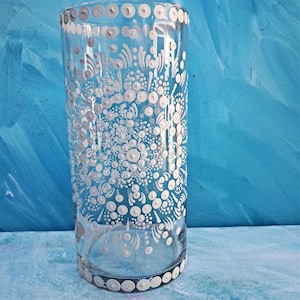 May include: A clear glass cylinder vase with a white dotted and floral design. The vase has a white dotted border at the top and bottom. The background is a textured blue.