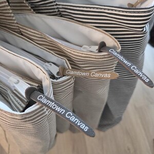 May include: Close-up of several canvas tote bags with horizontal stripes in various colors, including black and white, and brown and white. Each bag has a zipper and a leather tag that reads "Clamtown Canvas."