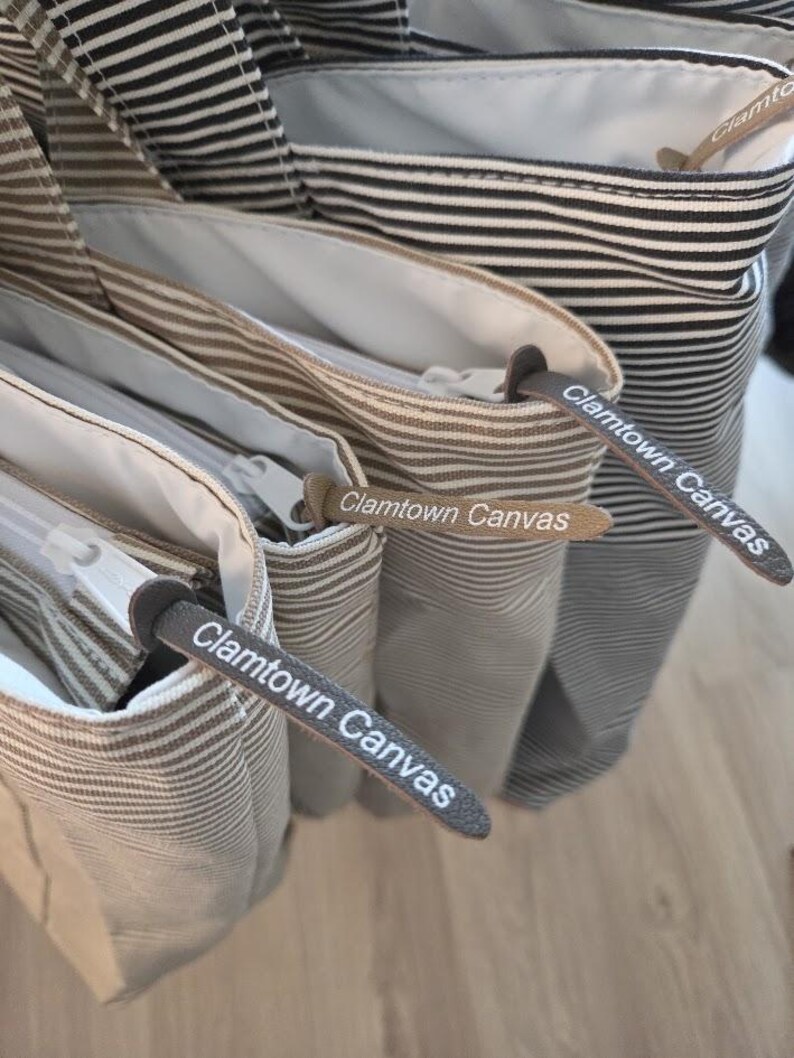 May include: Close-up of several canvas tote bags with horizontal stripes in various colors, including black and white, and brown and white. Each bag has a zipper and a leather tag that reads "Clamtown Canvas."