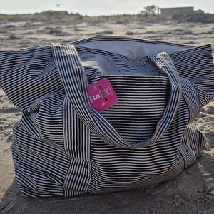 May include: A large tote bag with black and white horizontal stripes rests on a sandy beach. The bag has two handles and a pocket. A pink tag with the text "SAT" and the number "5" is attached to the bag.