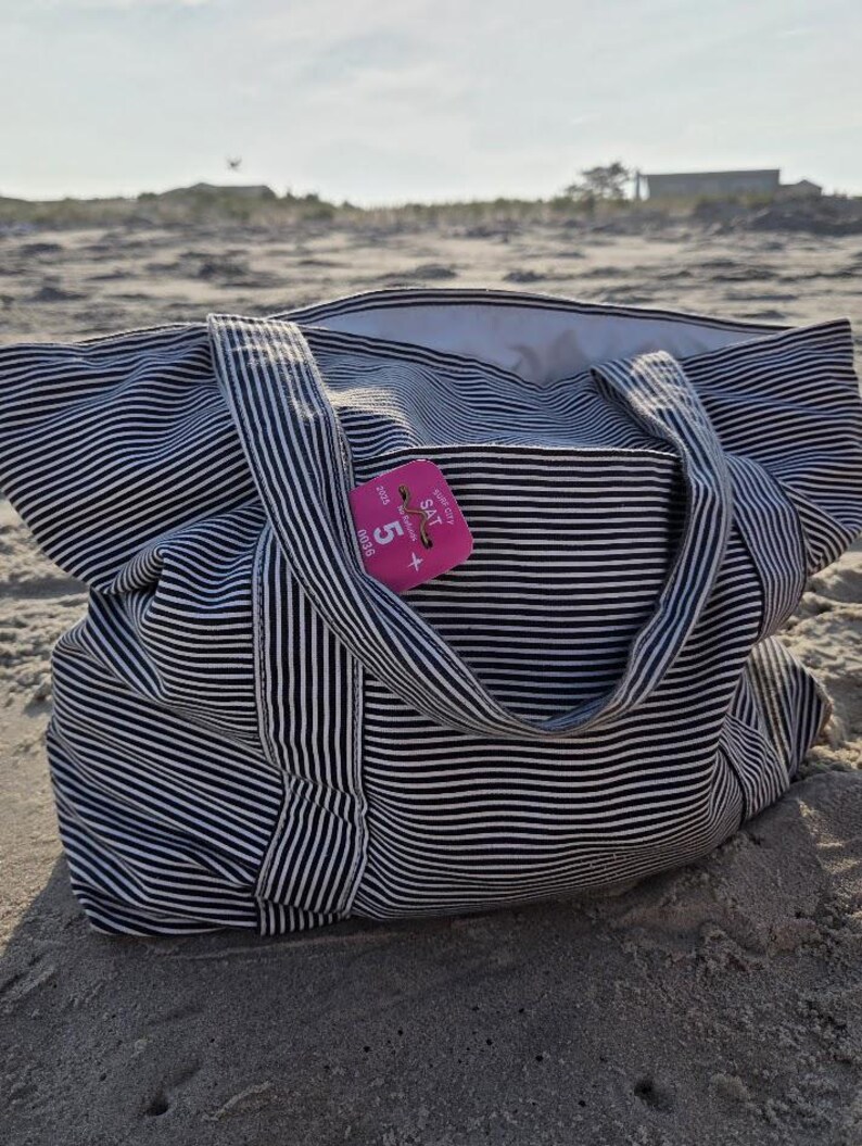 May include: A large tote bag with black and white horizontal stripes rests on a sandy beach. The bag has two handles and a pocket. A pink tag with the text "SAT" and the number "5" is attached to the bag.