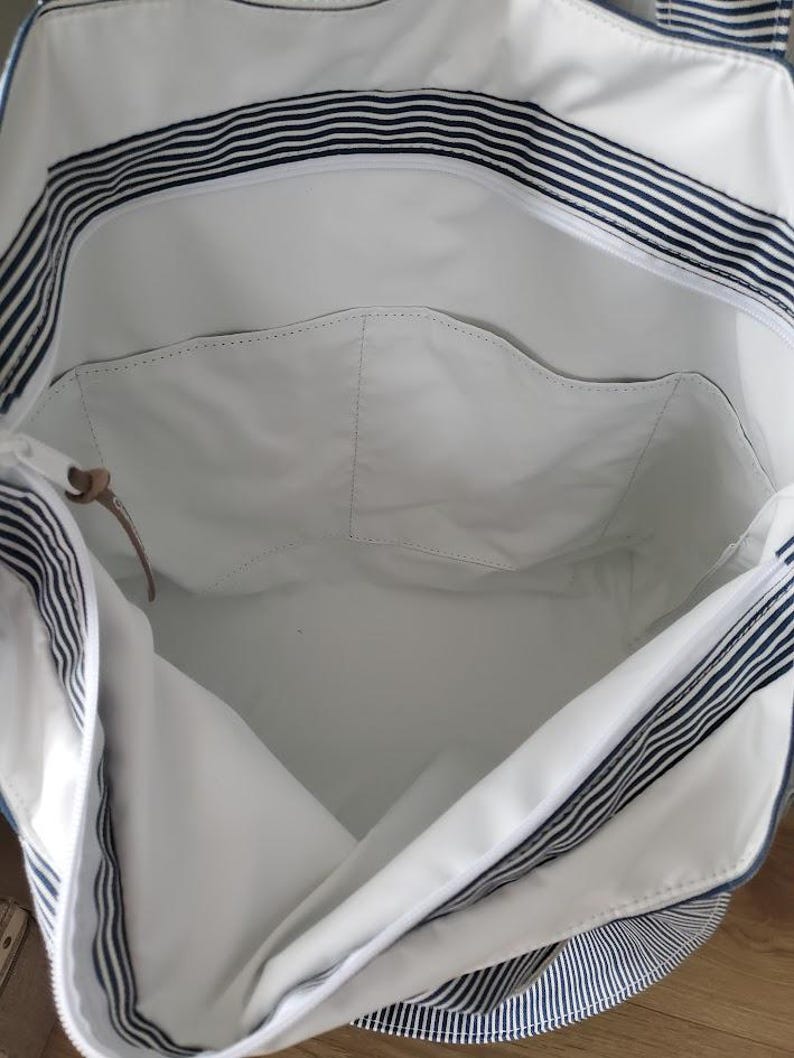 May include: White tote bag with navy blue stripes. The bag has a large interior with a zippered pocket and a small leather accent. The exterior features a striped pattern, and the interior is a solid white color.
