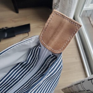 May include: Close-up of a striped tote bag with a light brown leather handle. The bag features navy blue and white stripes. The handle is rectangular and stitched with white thread.