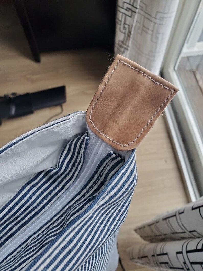May include: Close-up of a striped tote bag with a light brown leather handle. The bag features navy blue and white stripes. The handle is rectangular and stitched with white thread.