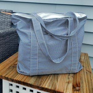May include: A large tote bag with blue and white horizontal stripes. The bag has two handles and a white interior lining. The bag is sitting on a wooden surface.