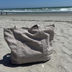 May include: A beige and white striped tote bag with sturdy handles sits on a sandy beach. The bag is made of a durable fabric and is designed for carrying items. The background shows the ocean and a clear blue sky.