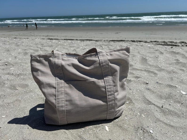 May include: A beige and white striped tote bag with sturdy handles sits on a sandy beach. The bag is made of a durable fabric and is designed for carrying items. The background shows the ocean and a clear blue sky.