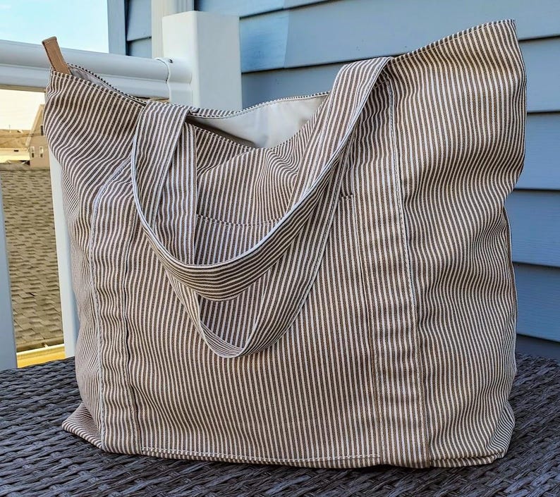 May include: A large tote bag with a brown and white striped pattern. The bag has a rectangular shape with a wide opening and two long straps. The bag is made of a soft fabric and has a small pocket on the front. The bag has a leather accent on the strap.