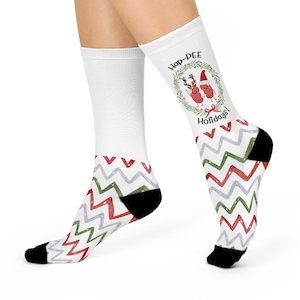 May include: White socks with a red, green, and gray zig-zag pattern and black heels and toes. The socks feature the text "Hap-PEE Holidays!" and a cartoon illustration of two red figures with reindeer antlers and a Santa hat.