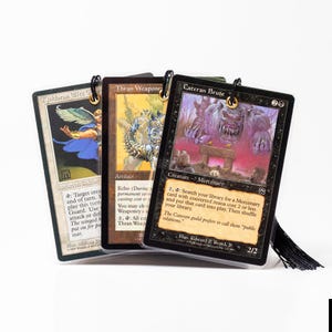 May include: Three collectible trading cards with protective sleeves. The cards feature fantasy art and text, including "Kjeldoran Elite," "Thran Weaponry," and "Cateran Brute." Each card has a black tassel attached at the top.