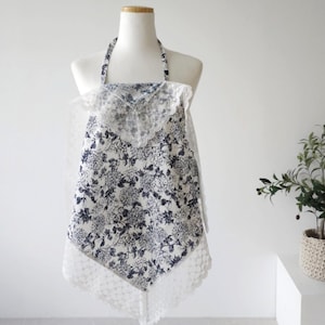 May include: A white apron with a blue floral print and white lace trim. The apron has a halter neck style and is tied at the back.