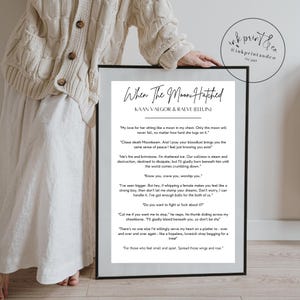 May include: A framed print with the title "When The Moon Hatched" and the names Kaan Vaegor & Raeve (Elluin). The print features a black frame and white text on a cream background. The image is held by a person wearing a cream-colored sweater.