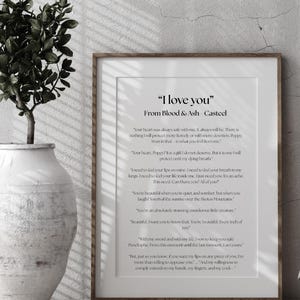 May include: A framed print with the text "I love you" from Blood & Ash Casteel. The print is in a wooden frame and is displayed against a neutral wall. A decorative vase with greenery is in the foreground.