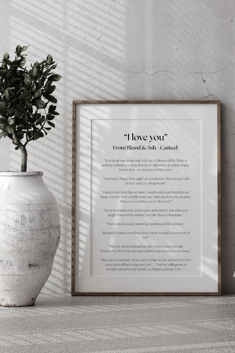 May include: A framed print with the text "I love you" from Blood & Ash Casteel. The print is in a wooden frame and is displayed against a neutral wall. A decorative vase with greenery is in the foreground.