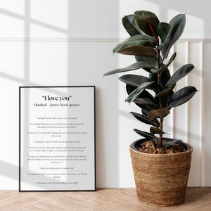 Puede incluir: Un marco negro con el texto "I love you" y citas de James Hook, junto a una gran planta de caucho en una cesta tejida. La planta tiene hojas grandes de color verde oscuro y un tallo rojizo.