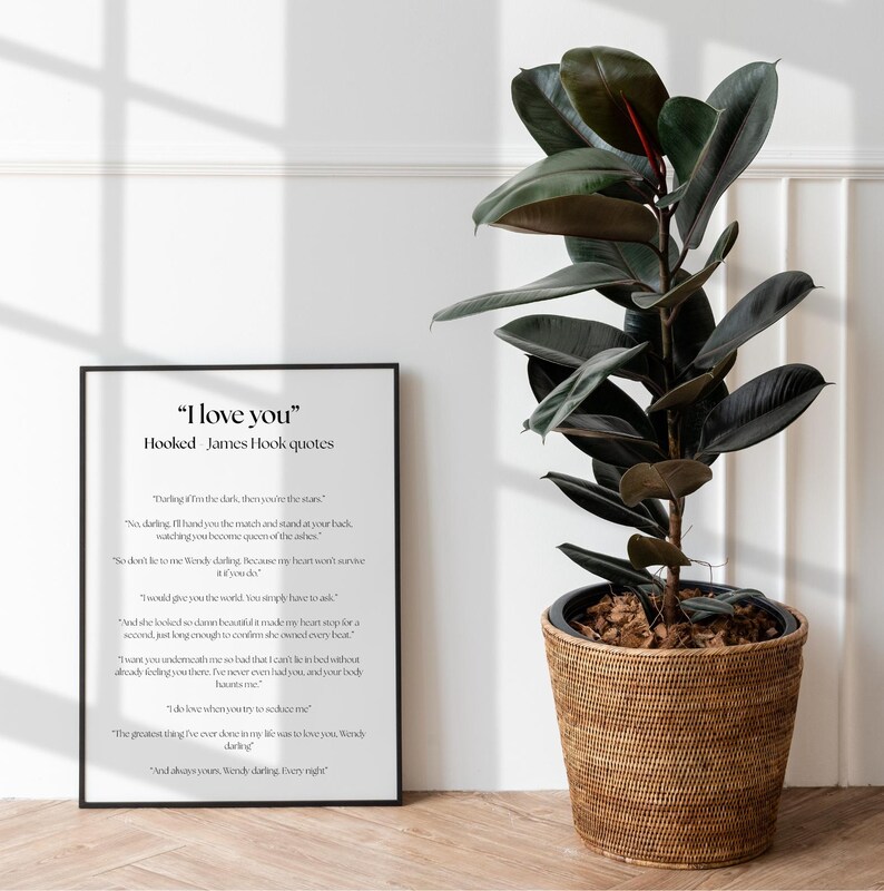 Puede incluir: Un marco negro con el texto "I love you" y citas de James Hook, junto a una gran planta de caucho en una cesta tejida. La planta tiene hojas grandes de color verde oscuro y un tallo rojizo.