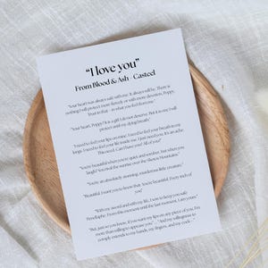 May include: A printed card with the text "I love you" from Blood & Ash Casteel, resting on a wooden plate. The card features romantic text in a serif font. The background includes a white cloth and dried flowers.