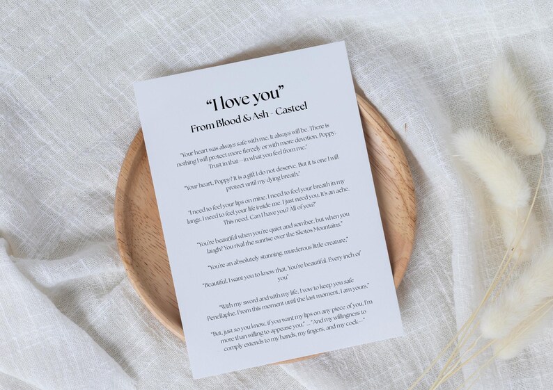May include: A printed card with the text "I love you" from Blood & Ash Casteel, resting on a wooden plate. The card features romantic text in a serif font. The background includes a white cloth and dried flowers.