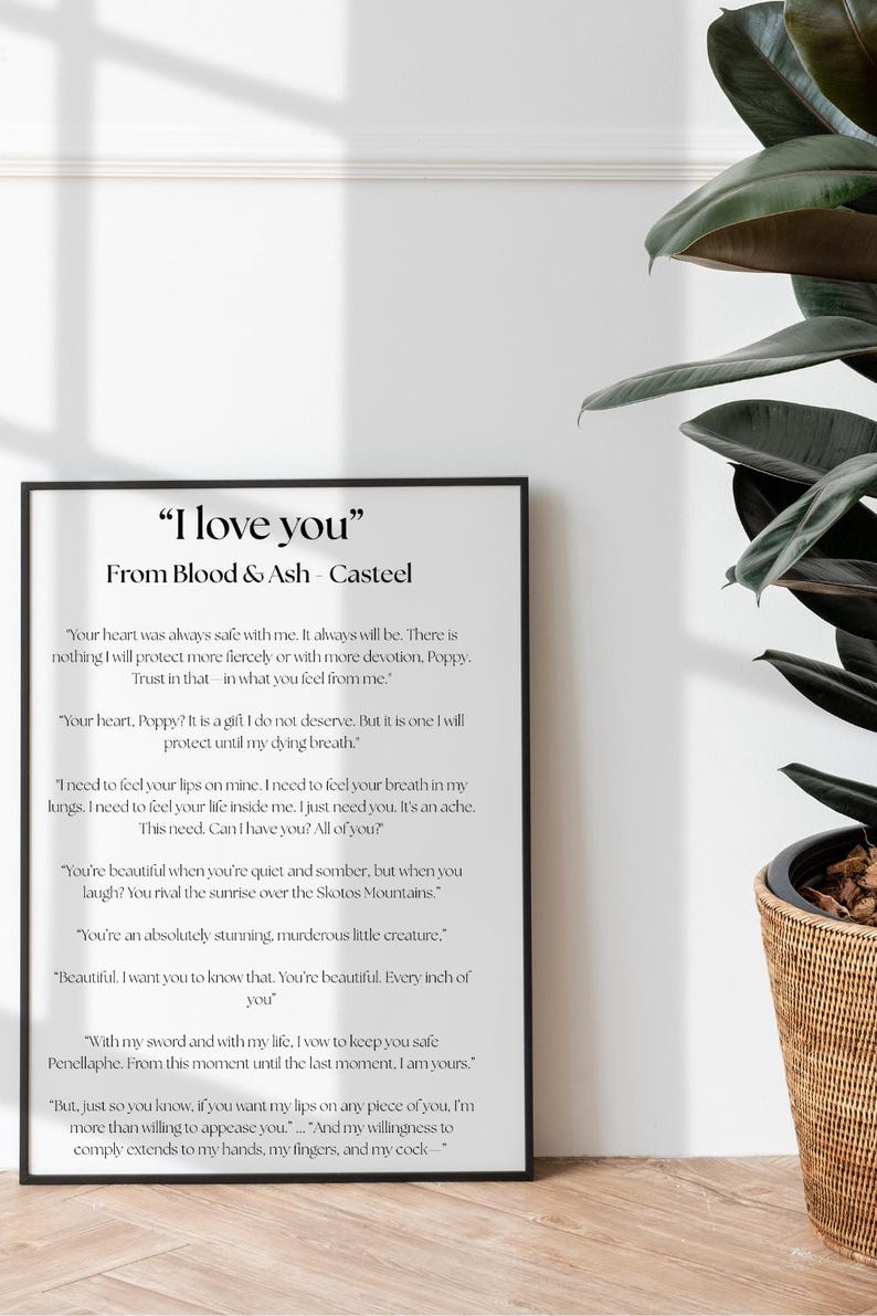May include: A framed print with the text "I love you" from Blood & Ash - Casteel. The black framed print is displayed against a white wall, with a large green plant in a woven basket to the right.