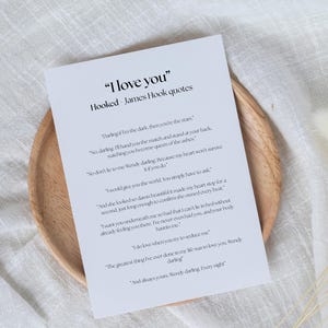 Puede incluir: Un papel blanco con el texto "I love you" y citas de James Hook, apoyado en un plato de madera. El papel tiene texto negro y est&aacute; rodeado por un fondo blanco y flores secas.