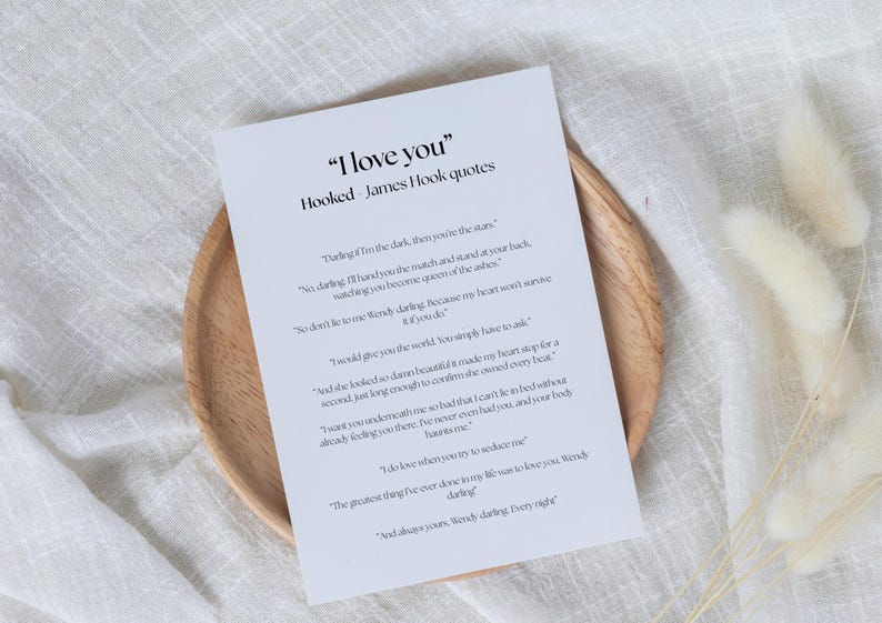 Puede incluir: Un papel blanco con el texto "I love you" y citas de James Hook, apoyado en un plato de madera. El papel tiene texto negro y est&aacute; rodeado por un fondo blanco y flores secas.