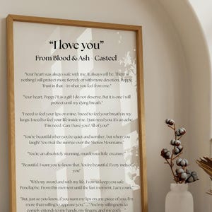 May include: A framed print with the text "I love you" from Blood & Ash - Casteel. The print has a light wood frame and features romantic quotes. A white vase with cotton stems sits to the right.