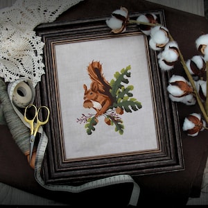 May include: A framed cross-stitch artwork featuring a brown squirrel holding acorns and perched on a green branch. The artwork is in a dark brown wooden frame. Cotton branches, lace, scissors, and a measuring tape are arranged around the frame.