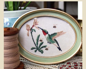May include: An oval framed cross-stitch design featuring a hummingbird and flowers, with a green and gold frame. The design is set against a white background. The text "The Hum Of The Garden" is at the bottom, with the designer's name at the top.