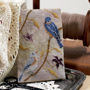 May include: A cross-stitch design featuring two bluebirds perched on a branch with yellow leaves and purple flowers. The artwork is displayed on a light beige fabric. A wooden spool of thread and a decorative lace cloth are in the background.