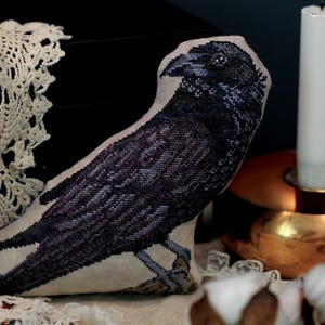 May include: A stuffed fabric ornament shaped like a crow, featuring detailed cross-stitch embroidery in shades of black, dark purple, and grey on a light beige linen background. The bird is depicted in profile, showcasing intricate feather textures.