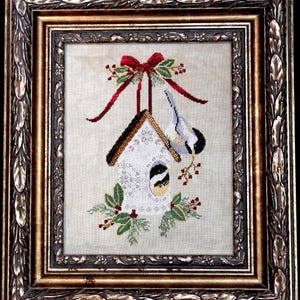 May include: Framed cross-stitch artwork featuring a white birdhouse with a black and gold roof, adorned with a red bow and sprigs of holly. A small bird perches on a branch near the birdhouse. The frame is ornate with gold and brown tones.