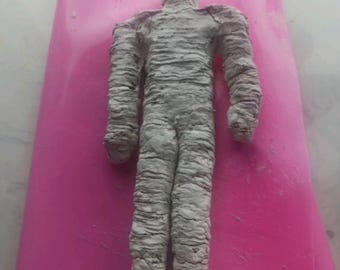 May include: A white clay sculpture of a person wrapped in bandages, resembling a mummy. The sculpture is standing on a pink surface.