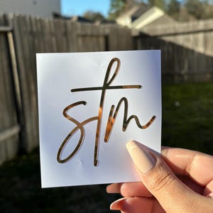 May include: Gold foil sticker with the word "stm" written in cursive script on a white background.