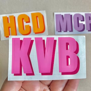 May include: Three stickers with different letters and colors. The first sticker is orange and says "HCD". The second sticker is purple and says "MCP". The third sticker is pink and says "KVB".