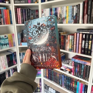 May include: A hardcover book titled "Forgotten Ashes" by Kathryn Covens, held in front of a bookshelf. The cover features a dark, ornate design with a crescent moon and the title in a stylized font. The background is a collection of various books.