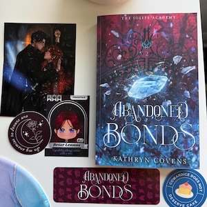 May include: A collection of items including a book titled "Abandoned Bonds" by Kathryn Covens, stickers, and a print. The book cover features a blue crystal and the title in white. Stickers include a round one with text "Enemies to lovers era" and a cartoon character.
