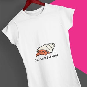 May include: White t-shirt featuring a cartoon hermit crab with an angry expression inside a shell. The text "Cute Shell. Bad Mood" is printed below the graphic. The shirt is hanging on a wooden hanger.