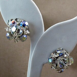 Puede incluir: Un par de pendientes de tono plateado con racimos de cristales transparentes. Los pendientes se muestran en un soporte blanco en forma de hoja.